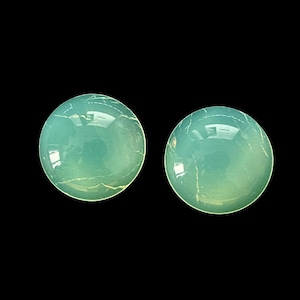 Puede incluir: Dos piedras preciosas redondas, de color verde azulado claro, con un aspecto pulido y translúcido. Cada piedra tiene sutiles vetas internas y un acabado brillante, sobre un fondo negro. Adecuado para la fabricación de joyas.
