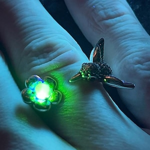 Puede incluir: Dos anillos, uno con forma de flor con un centro brillante, el otro con forma de colibrí. Los anillos son plateados y parecen llevarse en un dedo. El anillo de la flor tiene un brillo verde y el anillo del colibrí tiene alas detalladas.