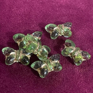 Puede incluir: Cuatro figuras de mariposas de cristal verde con detalles oscuros. Las mariposas están dispuestas sobre una superficie de color púrpura oscuro. El cristal es translúcido, permitiendo el paso de la luz.
