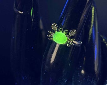 May include: A silver ring with a crab design. The crab has a glowing green center stone and small clear accent stones. The ring is shown on a black surface.