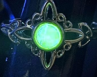 May include: A silver-toned ring with an ornate, symmetrical design. The ring features a central, glowing green stone, surrounded by a detailed filigree pattern. The ring is set against a dark background, highlighting the bright green gemstone.