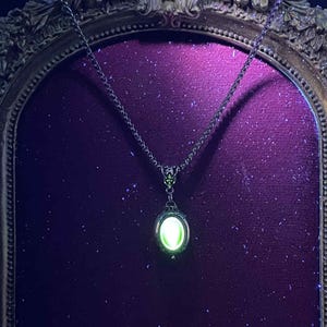 Op de afbeelding: Een zilverkleurige ketting met een gloeiende, ovaalvormige hanger. De hanger is gezet in een sierlijk frame en straalt een heldergroen licht uit. De ketting hangt tegen een donkerpaarse achtergrond, wat een opvallend contrast creëert.