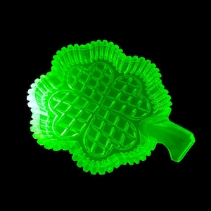 Puede incluir: Un plato verde brillante en forma de trébol de cuatro hojas, con un interior texturizado con diseño de diamantes. El plato tiene un borde festoneado y un pequeño asa. El diseño del trébol es un símbolo de buena suerte, a menudo asociado con el Día de San Patricio.