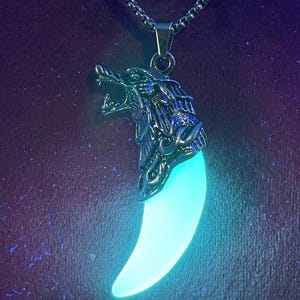 May include: A silver-colored pendant necklace featuring a wolf head design above a glowing, light blue tooth-shaped charm. The chain is a silver-colored, linked design. The background is a dark purple color.