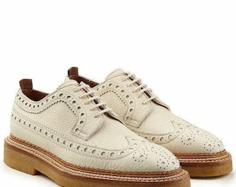 May include: A pair of off-white leather brogue shoes with decorative perforations and a textured appearance. The shoes feature a light brown sole and laces. The footwear is designed for everyday wear.