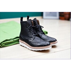 May include: A pair of black leather brogue boots with white soles and brown accents. The boots have a classic lace-up design and decorative perforations. The boots are placed on a light wood surface.