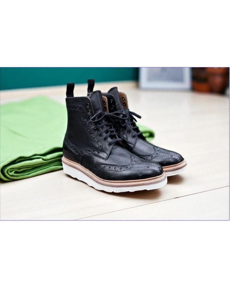 May include: A pair of black leather brogue boots with white soles and brown accents. The boots have a classic lace-up design and decorative perforations. The boots are placed on a light wood surface.