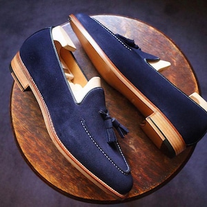 May include: A pair of navy blue suede loafers with a tassel detail and white stitching. The shoes have a light brown sole and heel, and are displayed on a wooden surface. The shoes are new and unworn.