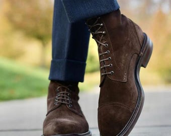 May include: Brown suede ankle boots with a lace-up design. The boots have a rounded toe and a dark brown sole. The image shows the boots paired with dark blue denim jeans.