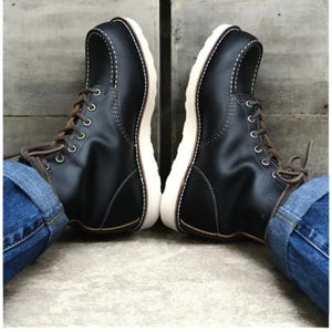 Puede incluir: Un par de botas de cuero negro con puntera estilo mocasín con costuras blancas en contraste, cordones marrones, ojales plateados y una suela gruesa de goma blanca. Las botas se llevan con vaqueros azules con los bajos doblados en los tobillos.