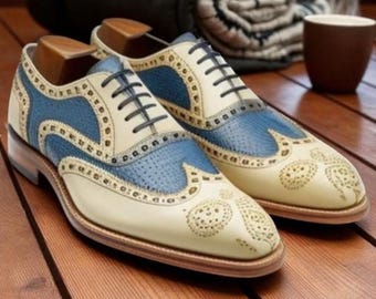 May include: Cream and blue leather brogue shoes with black laces. The shoes feature decorative perforations and a classic wingtip design. The shoes are displayed on a wooden surface.