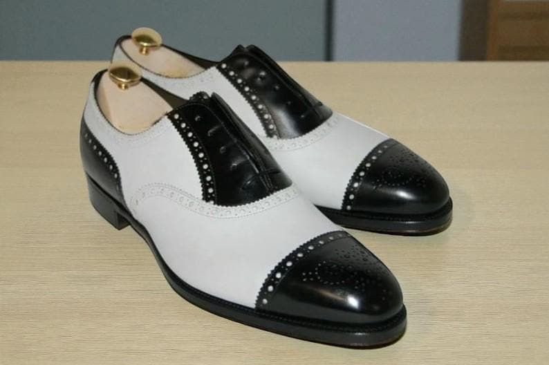 May include: A pair of two-tone, black and white leather dress shoes. The shoes feature a classic wingtip design with perforated detailing along the seams. The toe and heel caps are black, while the main body is white.