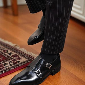 May include: Black leather double monk strap shoes with silver buckles. The shoes have a glossy finish and are worn with black socks and pinstripe pants. The shoes are on a wooden floor.