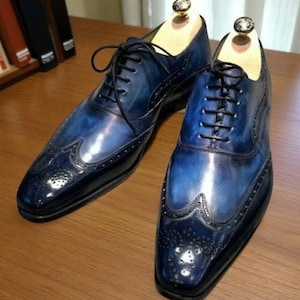 May include: A pair of blue leather dress shoes with a wingtip design. The shoes have a glossy finish and black laces. Shoe trees are inserted in the shoes. The shoes are on a wooden surface.