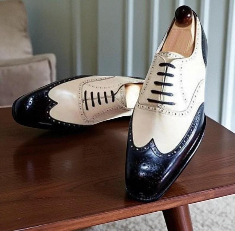 May include: A pair of two-tone wingtip dress shoes. The shoes feature a cream-colored upper with black accents on the toe and heel. Black laces and decorative perforations add detail. The shoes are displayed on a wooden surface.
