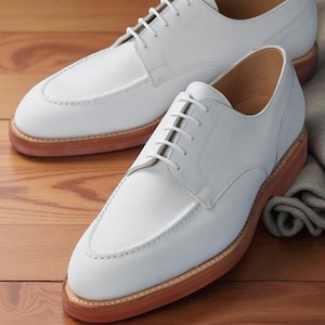 May include: A pair of white leather shoes with a classic design. The shoes have a light brown sole and white laces. The shoes are displayed on a wooden surface.