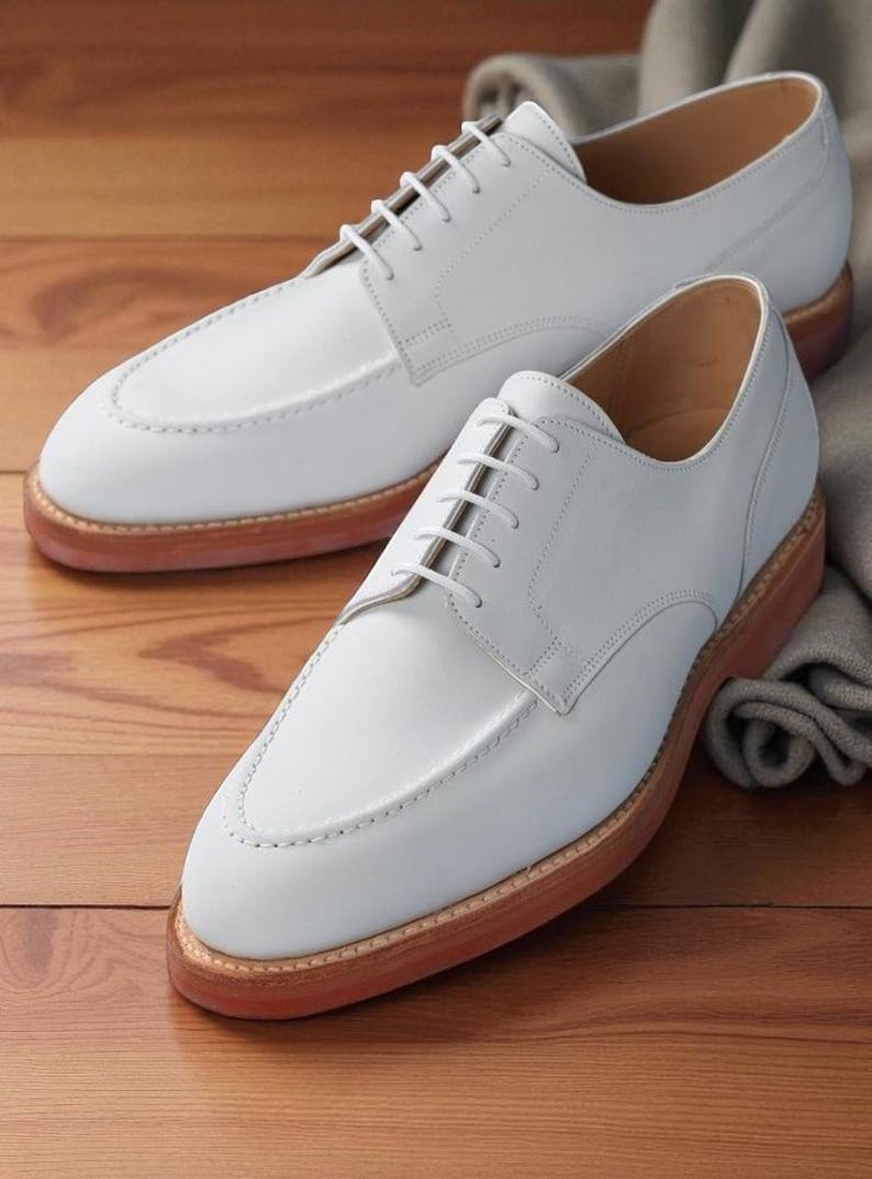May include: A pair of white leather shoes with a classic design. The shoes have a light brown sole and white laces. The shoes are displayed on a wooden surface.