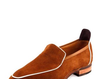 Puede incluir: Un mocasín de ante marrón con ribete blanco y tacón pequeño. El zapato tiene punta redondeada y un detalle marrón oscuro en el talón. La suela es de color marrón claro.