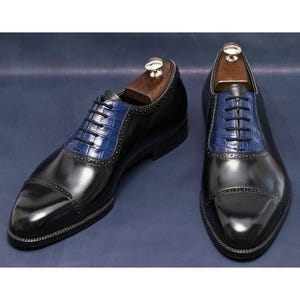 May include: A pair of black leather dress shoes with blue textured panels on the vamp and a cap toe design. The shoes have black laces and are displayed with wooden shoe trees.