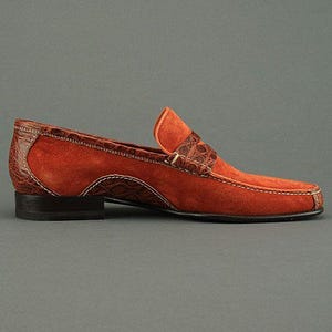 K&ouml;nnte beinhalten: Ein mokkafarbener Wildleder-Loafer mit braunen Kroko-Leder-Akzenten. Der Schuh hat eine eckige Spitze, einen niedrigen Absatz und wei&szlig;e N&auml;hte. Eine stilvolle Schuhoption.