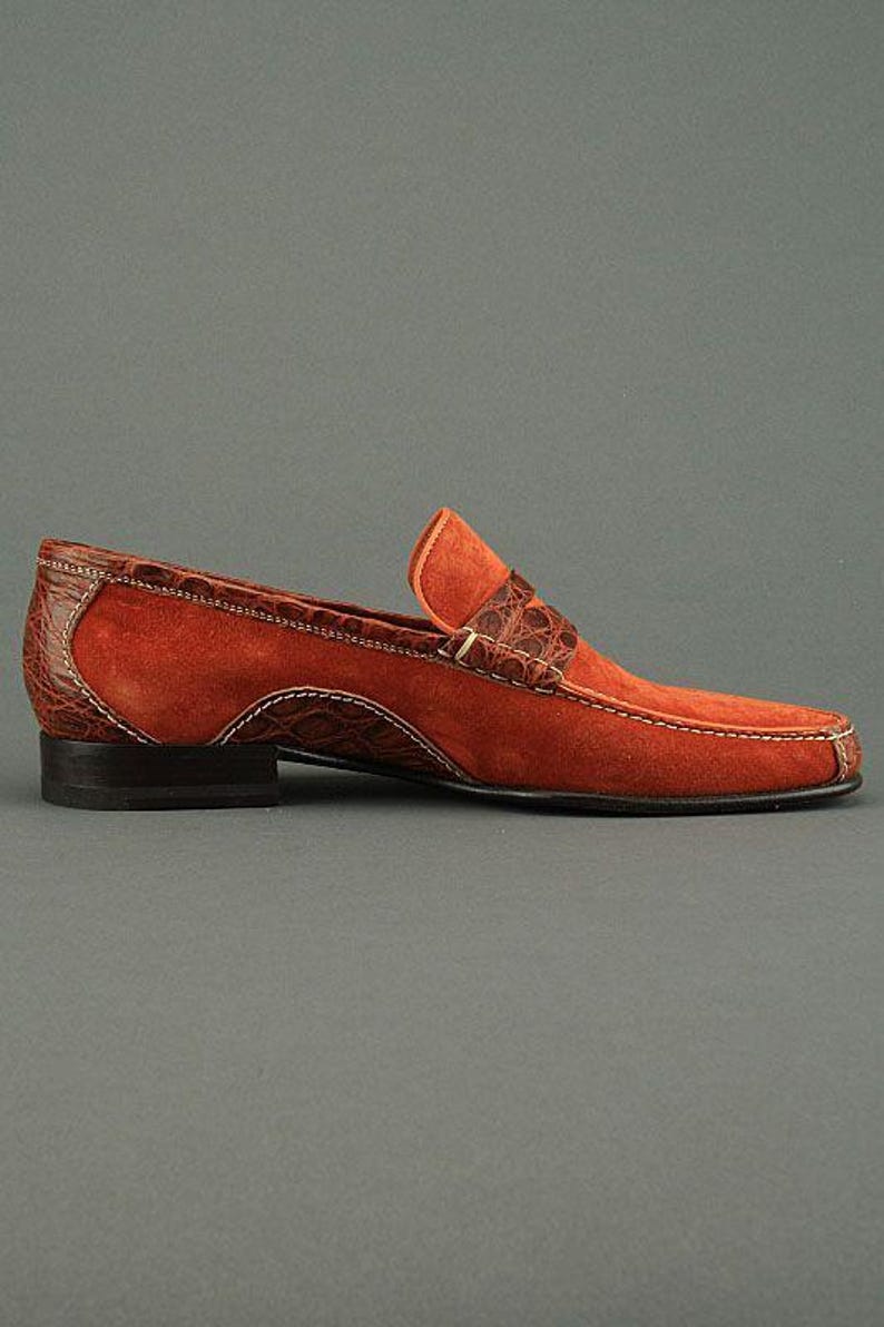 K&ouml;nnte beinhalten: Ein mokkafarbener Wildleder-Loafer mit braunen Kroko-Leder-Akzenten. Der Schuh hat eine eckige Spitze, einen niedrigen Absatz und wei&szlig;e N&auml;hte. Eine stilvolle Schuhoption.
