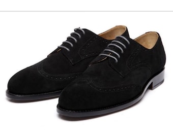 May include: A pair of black suede Oxford shoes with gray laces. The shoes have a classic wingtip design with decorative perforations along the seams. The soles are black.