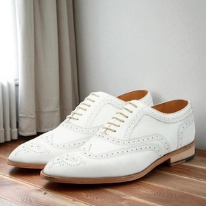 May include: A pair of white leather brogue shoes with decorative perforations and light-colored laces. The shoes have a classic design with a low heel and are displayed on a wooden surface.