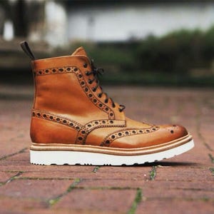 May include: A brown leather boot with brogue detailing and a white sole. The boot has a lace-up closure and a pull tab at the back. The boot is made of leather and has a classic design.