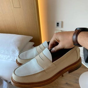 K&ouml;nnte beinhalten: Ein Paar cremefarbene Leder-Loafer mit hellbraunem Akzent am Oberteil und einer braunen Sohle. Die Schuhe im klassischen Penny-Loafer-Design werden auf einer Holzoberfl&auml;che pr&auml;sentiert. Ca. 25 cm lang.