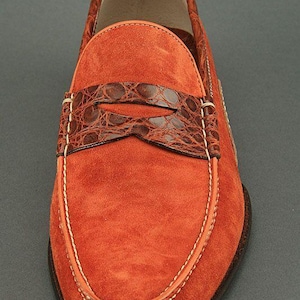 K&ouml;nnte beinhalten: Orangener Wildleder-Loafer mit einem braunen Riemen aus Krokodilleder-Optik &uuml;ber dem Spann. Der Schuh hat wei&szlig;e N&auml;hte und eine dunkelbraune Sohle. Eine stilvolle Schuhoption.