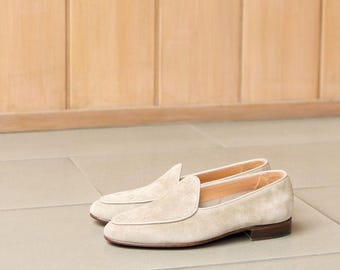 Puede incluir: Un par de mocasines de ante beige. Estos zapatos clásicos presentan una puntera redondeada, un tacón bajo y un ribete blanco. Los mocasines se exhiben sobre una superficie de baldosas de color claro.