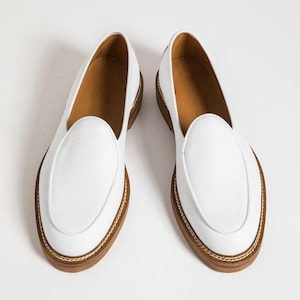 Puede incluir: Un par de mocasines de cuero blanco con punta redondeada y suela marr&oacute;n. Los zapatos tienen un dise&ntilde;o cl&aacute;sico con detalles cosidos alrededor de la suela. El interior del zapato es de color marr&oacute;n claro.
