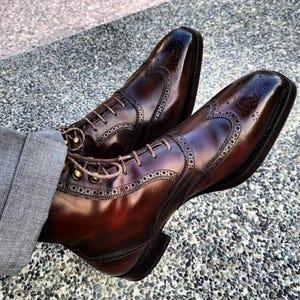 Könnte beinhalten: Ein Paar polierte, dunkelbraune Lederschuhe mit aufwendigen Brogue-Details. Die Schuhe haben ein klassisches Flügelspitzen-Design mit dekorativen Perforationen und einem Schnürverschluss. Das Leder hat eine glänzende Oberfläche.