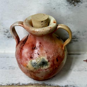 May include: A small, red and white ceramic pitcher with two handles and a cork stopper. The pitcher has a rustic, hand-painted look.