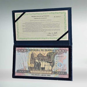 Puede incluir: Un billete conmemorativo de 2000 Piso de Filipinas enmarcado, presentado en un estuche azul oscuro. El billete presenta una escena histórica y el texto "REPUBLIKA NG PILIPINAS 2000". Un certificado de emisión es visible arriba.