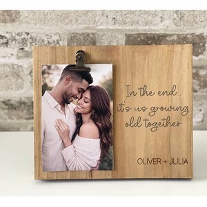 May include: A wooden photo frame with a picture of a couple, held by a metal clip. The frame has the text "In the end it's us growing old together" and the names "OLIVER + JULIA" engraved on it.