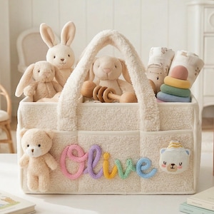 May include: A cream-colored baby tote bag filled with plush toys, including bunnies and a teddy bear. The name "Olive" is spelled out in colorful yarn on the front. The bag also contains baby blankets and a wooden stacking toy.