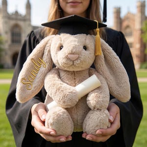 Puede incluir: Un conejito de peluche con birrete de graduación y borla dorada, sosteniendo un diploma enrollado. El conejo es de color marrón claro con largas orejas caídas y el nombre "Sandy" bordado en una oreja. El fondo muestra a una persona con toga de graduación.