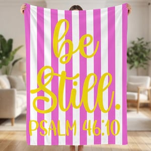 May include: A pink and white striped blanket with the words "be still" in yellow script. The text "Psalm 46:10" is below the script. The blanket is held up in front of a blurred background.