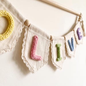 May include: A decorative wall banner featuring off-white canvas pennants trimmed with cream lace. Each pennant displays a single letter in a raised, knitted texture. The letters spell out the word LOVE in yellow, pink, green, and blue.