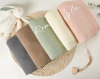 May include: Five knitted baby blankets in different colours, with the names "Lyla", "Rom", "Leo", "Ava", and "Mia" embroidered on them. The blankets are arranged in a circle on a woven mat.