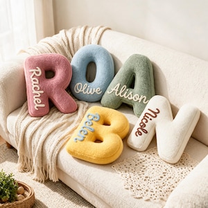 Pode incluir: Cinco almofadas de pelúcia em forma de letra nas cores rosa, azul, verde, amarelo e branco. Cada almofada apresenta um nome bordado em letra cursiva: Rachel, Olive, Alison, Belen e Nicole. As almofadas têm uma superfície texturizada e felpuda.