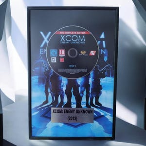 Può includere: Un display incorniciato con il disco del gioco per PC XCOM: Enemy Unknown The Complete Edition montato su una stampa grafica dai toni blu di soldati. Una targhetta argentata in basso riporta la scritta XCOM: ENEMY UNKNOWN (2012) in una cornice nera.
