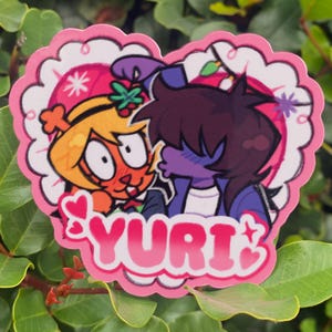 May include: A heart-shaped sticker featuring two cartoon characters in a romantic embrace. The sticker has a pink scalloped border and the word "YURI" in pink lettering. The characters are in shades of orange, purple, and blue.