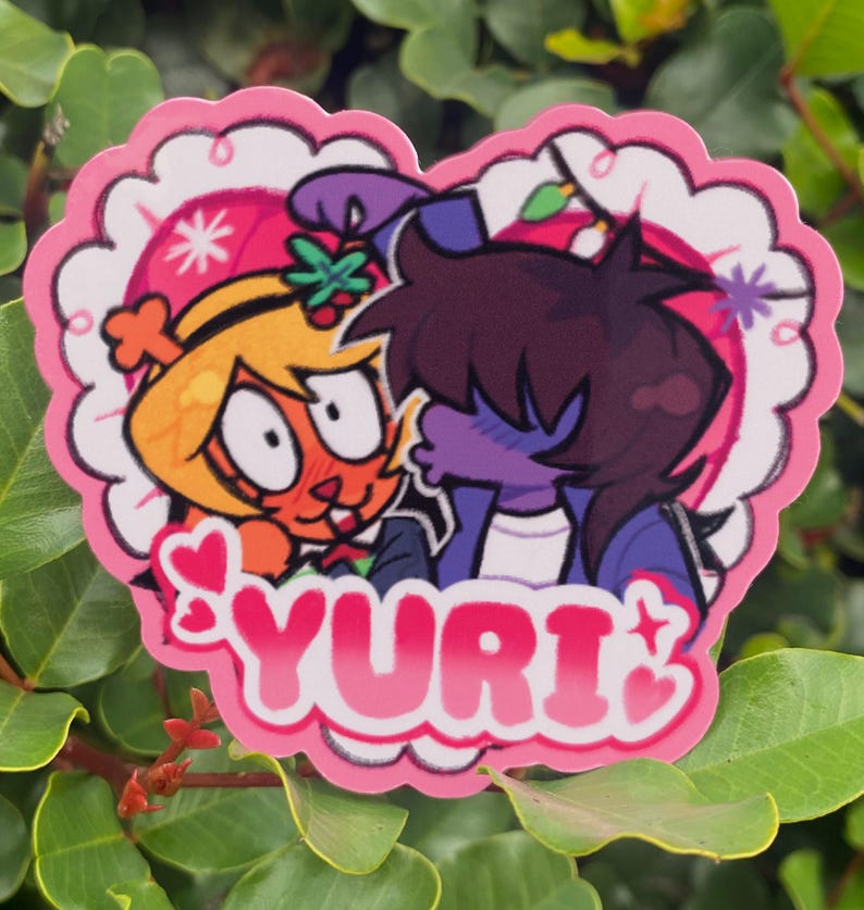 May include: A heart-shaped sticker featuring two cartoon characters in a romantic embrace. The sticker has a pink scalloped border and the word "YURI" in pink lettering. The characters are in shades of orange, purple, and blue.