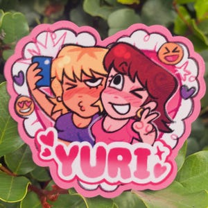 May include: A heart-shaped sticker featuring two cartoon women taking a selfie. One woman has blonde hair and the other has red hair. The sticker has pink accents, hearts, and the word "YURI".