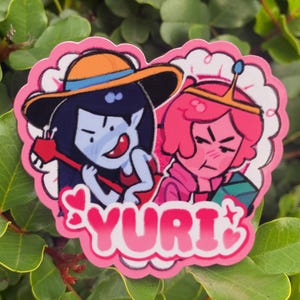 May include: A pink heart-shaped sticker featuring two cartoon characters. One character has blue skin, a hat, and is holding a red instrument. The other character has pink skin and a crown. The word "YURI" is written in pink letters.