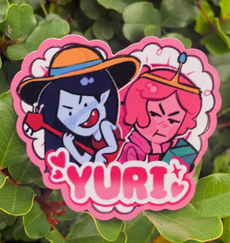 May include: A pink heart-shaped sticker featuring two cartoon characters. One character has blue skin, a hat, and is holding a red instrument. The other character has pink skin and a crown. The word "YURI" is written in pink letters.