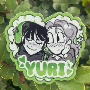 May include: A green sticker featuring two anime-style characters with musical notes. The sticker has a heart-shaped border with the word "YURI" in green. One character has black hair and the other has gray hair.