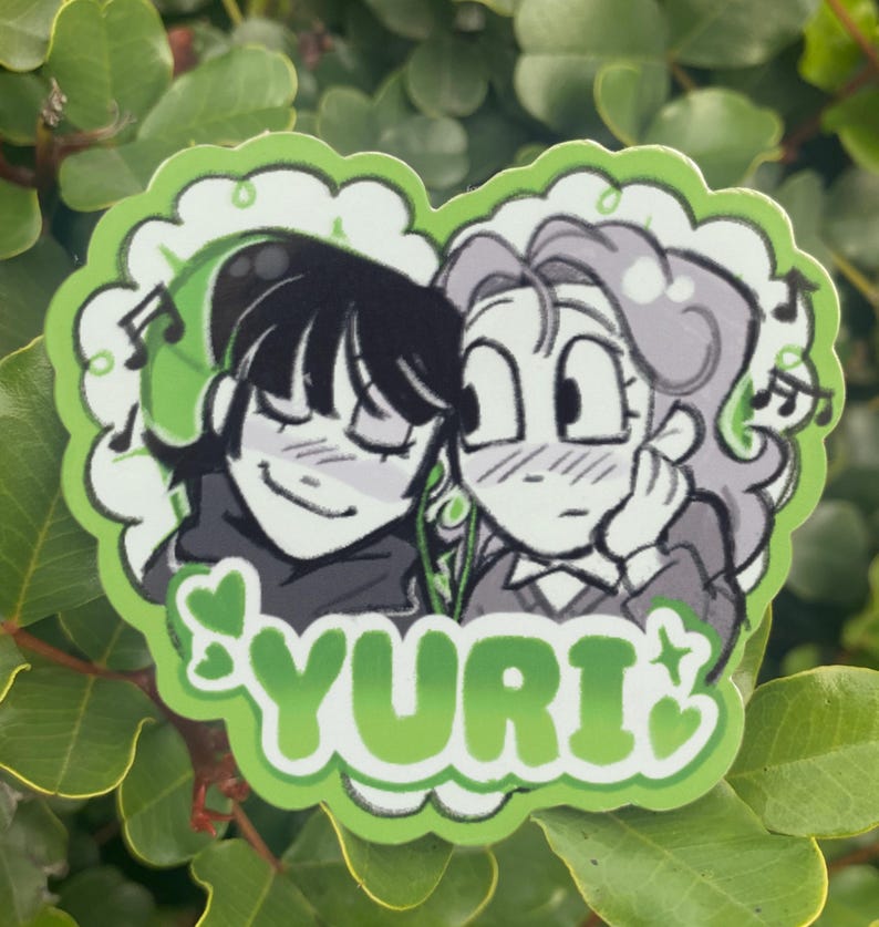 May include: A green sticker featuring two anime-style characters with musical notes. The sticker has a heart-shaped border with the word "YURI" in green. One character has black hair and the other has gray hair.